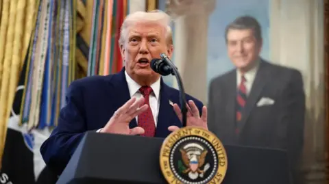 U.S. President Donald Trump speaks at a lectern in a navy suit with a red tie in front of a painting and flags