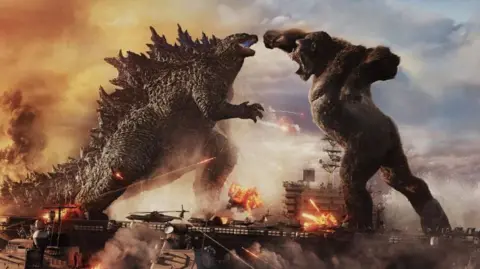 A still from the Godzilla vs Kong movie. The two titular monsters engage in battle while standing on military battle ships.